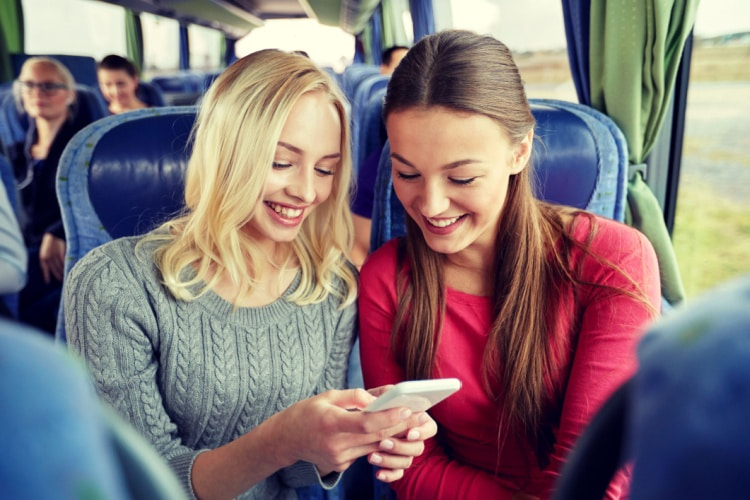 two students look at a cell phone while sitting together on a charter bus 